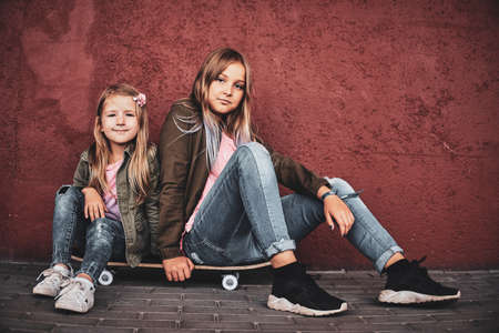 Two pretty little girls are chilling on the street while sitting on the skateboard.の写真素材