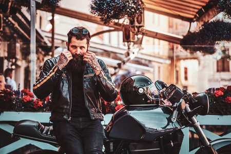 Handssome serious biker with beard and tattooes is sitting on his motorbike in busy city centre.の写真素材