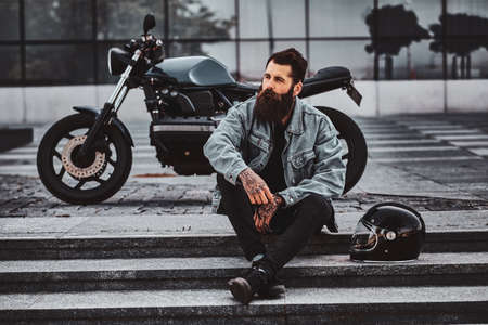 Brutal pensive man in denim jacket is sitting on the stairs close to his motorbike neaar glass building.の写真素材