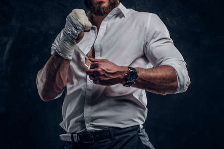 Bearded brutal man in white shirt is wearing protection on his fist before fight at dark studio.の写真素材