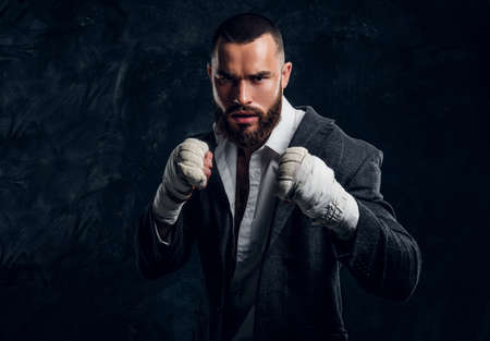 Angry bearded businessman in suit and protective kickboxing gloves is posing for photographer at dark photo studio.の写真素材