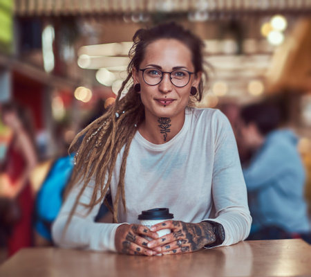 Beautiful cheerful girl with tattooes and dreadlocks is sitting at food court while drinking coffee.