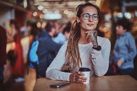Beautiful cheerful girl with tattooes and dreadlocks is sitting at food court while drinking coffee.の写真素材