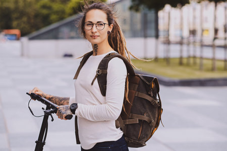 Beautiful trendy girl with dreadlocks is driving electro scooter on the street in cloudy summer day.