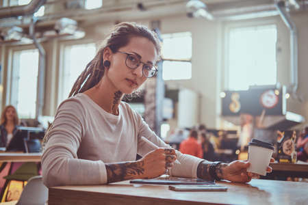 Styilish pensive girl is sketching on digital notepad while sitting at cafe with cup of coffee.の写真素材