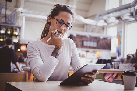 Pretty creative girl in glasses is drawing on digital notepad while sitting at cafeteria.