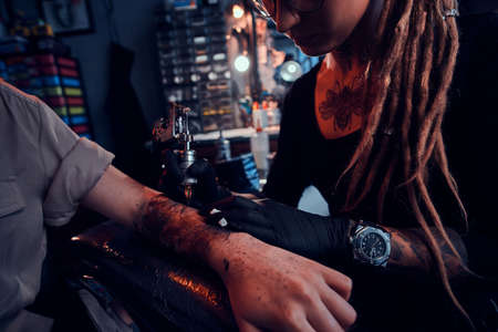 Young focused tattoo master is makining tattoo on clients hand at dark tattoo salon.の写真素材