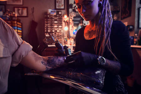 Creative tattoo master with dreadlocks is creating beautiful tattoo on womans hand at tattoo salon.の写真素材