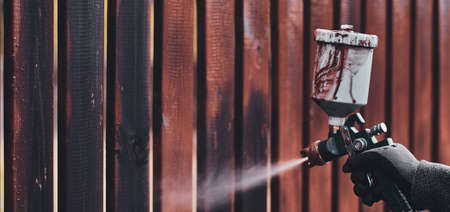 Process of old fence renovation by worker in protective gloves. He is usin airbrush, applying paint.の写真素材