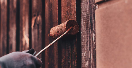 Old fences renovaiting process by worker. He apply brown paint with brush, wearing protective...