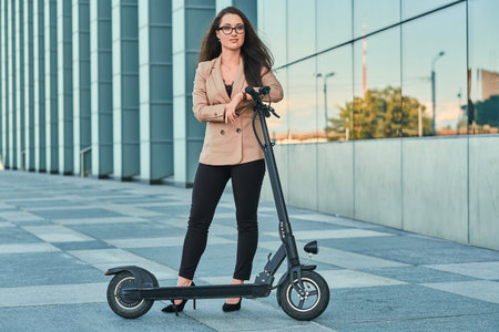Young woman in formal wear and glasses is riding electrical scooter from her work.