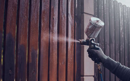 Process of old fence renovation by worker in protective gloves. He is usin airbrush, applying paint.の写真素材