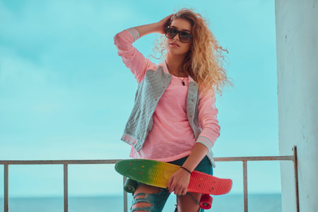 Young blond woman in sunglasses is posing for photographer while holding her skateboard.