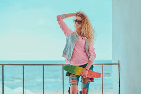 Young blond woman in sunglasses is posing for photographer while holding her skateboard.の写真素材