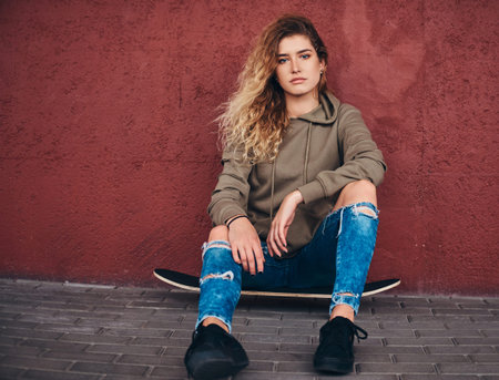 Beautiful young woman in ripped jeans is sitting on her own skateboard at the street.