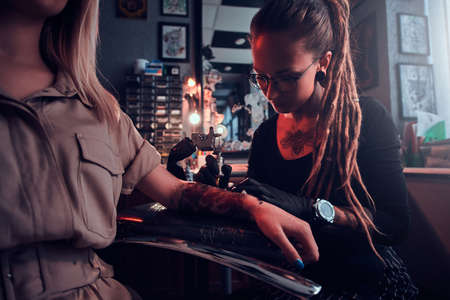 Young focused tattoo master is makining tattoo on clients hand at dark tattoo salon.の写真素材