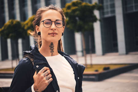 Nice young woman with piercing, glasses and tattoo on her neck is posing for photographer on the street.の写真素材
