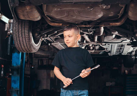 Little boy is standing under the car at auto workshop with wrench in his hand.の写真素材