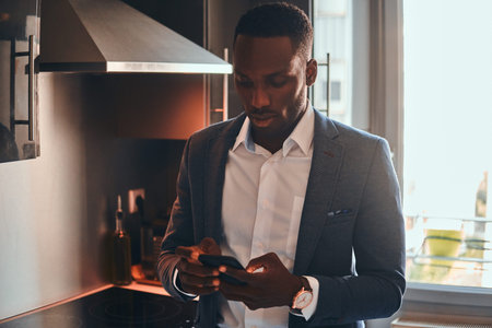 Young african man in white shirt is chatting by mobile phone at the kitchen.の写真素材