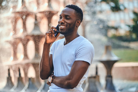 Happy smiling man is talking by mobile phone while chilling near fountain.