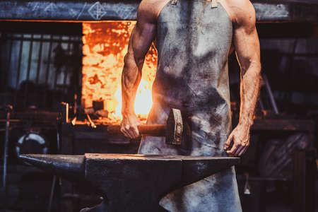 Muscular blacksmith in protective apron at his workshop with hammer in his hands. There are fire at background.の写真素材