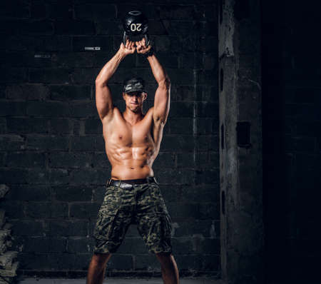 Focused shirtless man in cap is doing exercises with dumbbell at dark photo studio.の写真素材