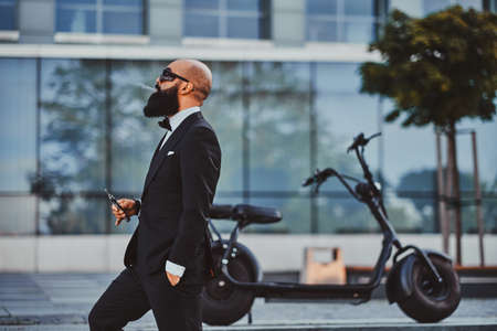 Attractive bald man in sunglasses is smoking, making nice vapour while standing nearhis electro bike.の写真素材