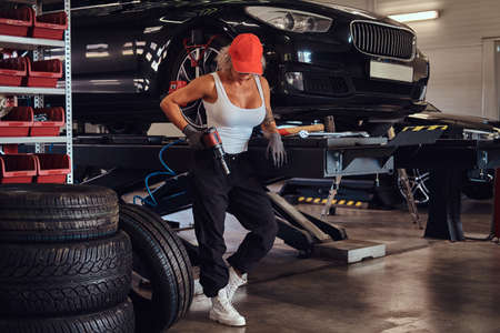 Sexy attractive woman worker in cap is sitting in workshop with pneumatic drill in hand.の写真素材