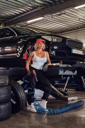 Sexy attractive woman worker in cap is sitting in workshop with pheumatic drill in hand.の写真素材