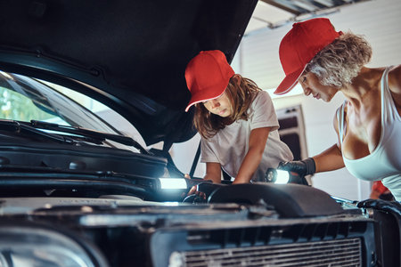 Little smiling girl is curious about cars engine at auto service with her mother, which is doing...