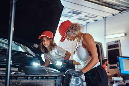 Mother is teaching her daughter how to fix broken cars engine at workshop.の写真素材