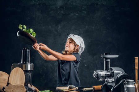 Pretty little girl is tossing vegetables on the pan at dark photo studio.の写真素材