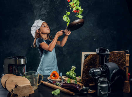 Pretty little girl is tossing vegetables on the pan at dark photo studio.の写真素材
