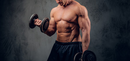 Attractive muscular bodybuilder is doing exercises with barbels and showing his muscules on the dark...