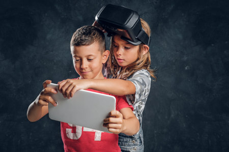 Little boy and girl are focused on tablet, girl has vr goggles on head.