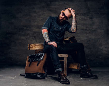 Pensive bearded man with tattooed arms is sitting at dark photo studio.の写真素材