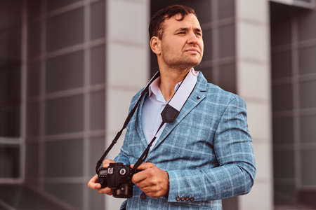 Attrative pensive photojournalist is standing with his digital photo camera near interesting building.の写真素材