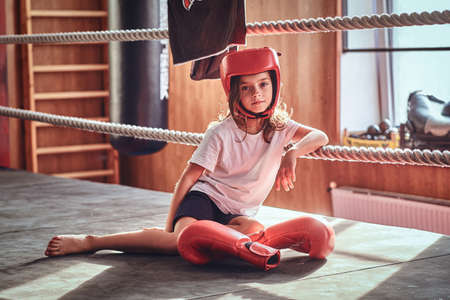 Beautiful kid girl is sitting on boxing ring wearing boxer uniform - gloves and helmet.の写真素材