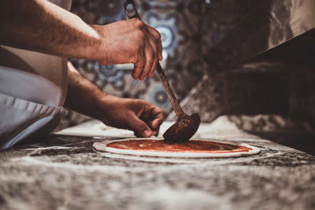Italian chef in uniform is adding tomato sauce for pizza at the kitchen.