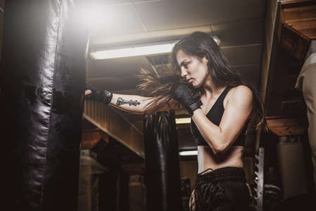 Skinny focused woman has a boxing training with punching bag at dark gym.の写真素材