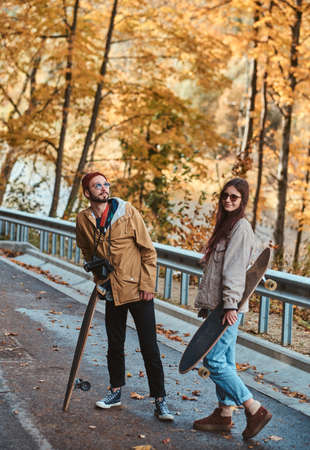 Beautiful romantic couple with longboards are walking in sunny autumn park.の写真素材