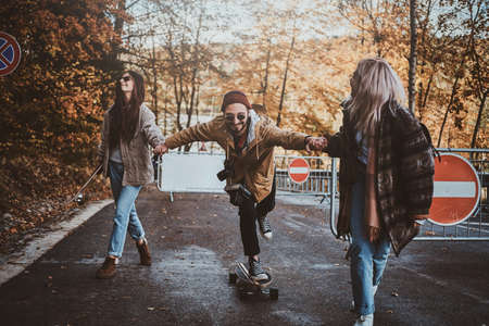 Two young women are teaching their friend how to ride skateboard while walking in autumn park.の写真素材