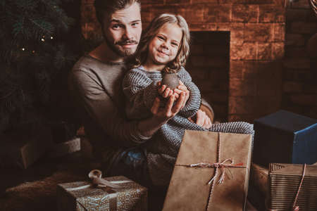 Cheerful little girl is holding christmas decoration ball while sitting with her father on the floor, surrounded with gifts.の写真素材