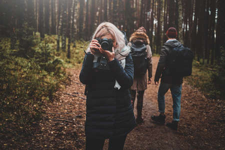 Young blond woman is making a photo with her camera while walking in forest with her friends.の写真素材