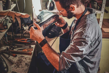 Diligent cobbler in glasses is working with pair of leather shoes at his workplace.の写真素材
