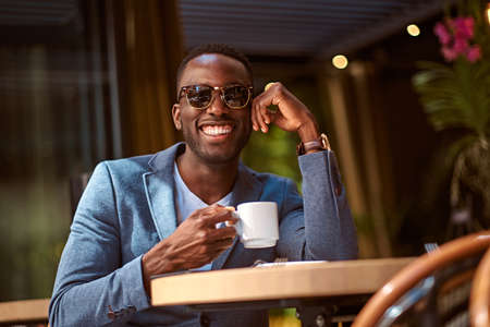 Smart american student is sitting at cafe while enjoying his cup of coffee.