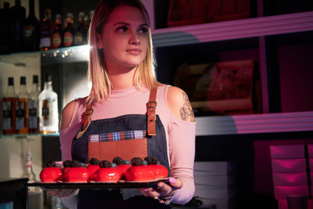 Young attractive woman is holding a tray with freshly prepared cakes at bakery store.