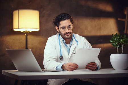 Young male doctor in glasses is holding documents in hands while working at night shift.