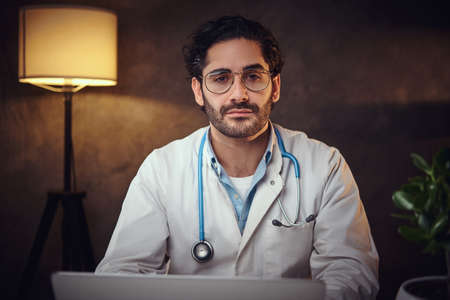 Handsome young doctor is working at late nightshift at his office using a laptop.の写真素材