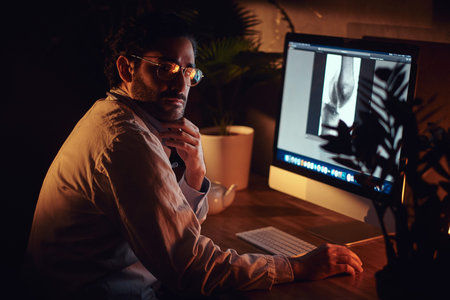 Pensive dilligent intern is working at late night shift at his office using a computer.